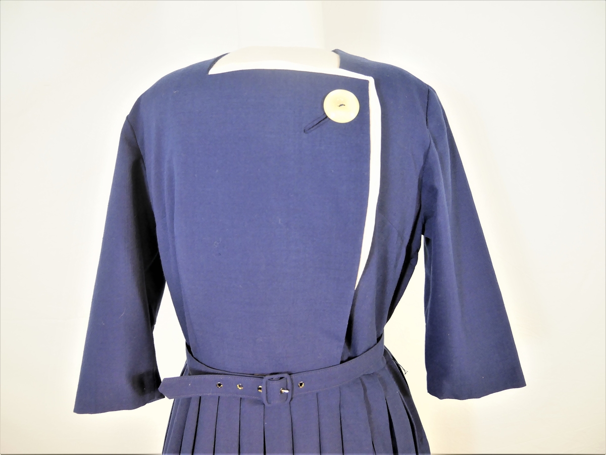 Mørkeblå kjole med plisseskjørt, og belte i livet. Ved halsen er det en hvit knapp og hvite detaljer. 