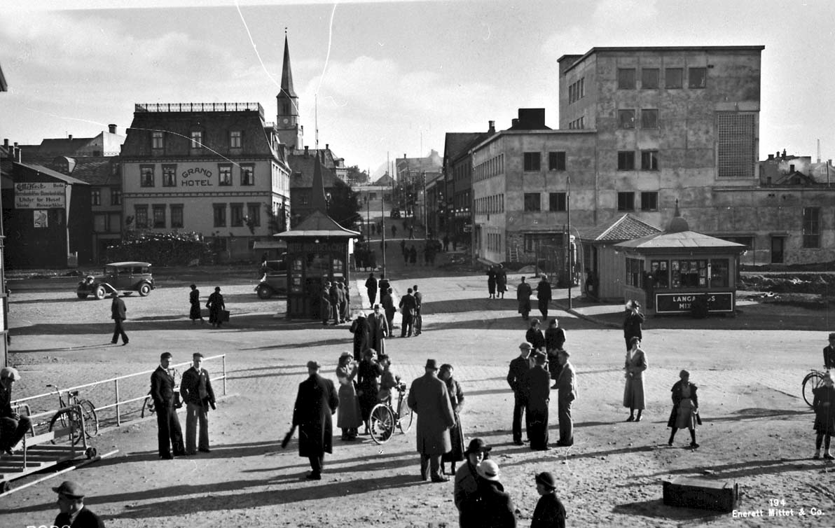 Tettsted - by. Åpen plass i forgrunnen med mange personer og noen biler. I bakgrunnen sees Grand Hotel og et kirketårn.
Bilde tatt i 1936