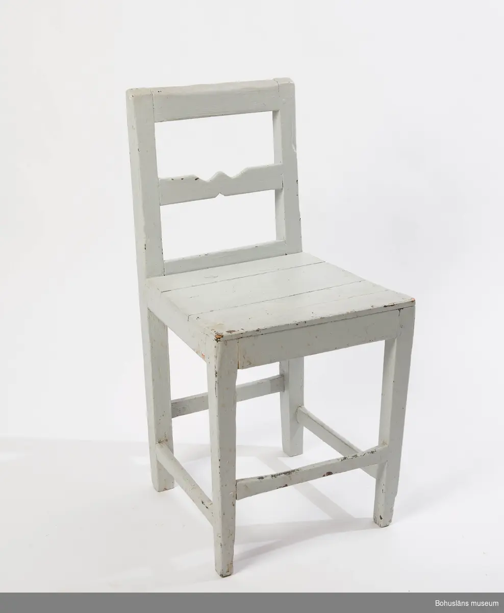 Vitmålad enkel stol med slät sits och nertill fyra tvärslåar på olika höjd.
Ryggstöd med sk. "krusabete", en dekorativ tilltäljd utsirning på mittslån.