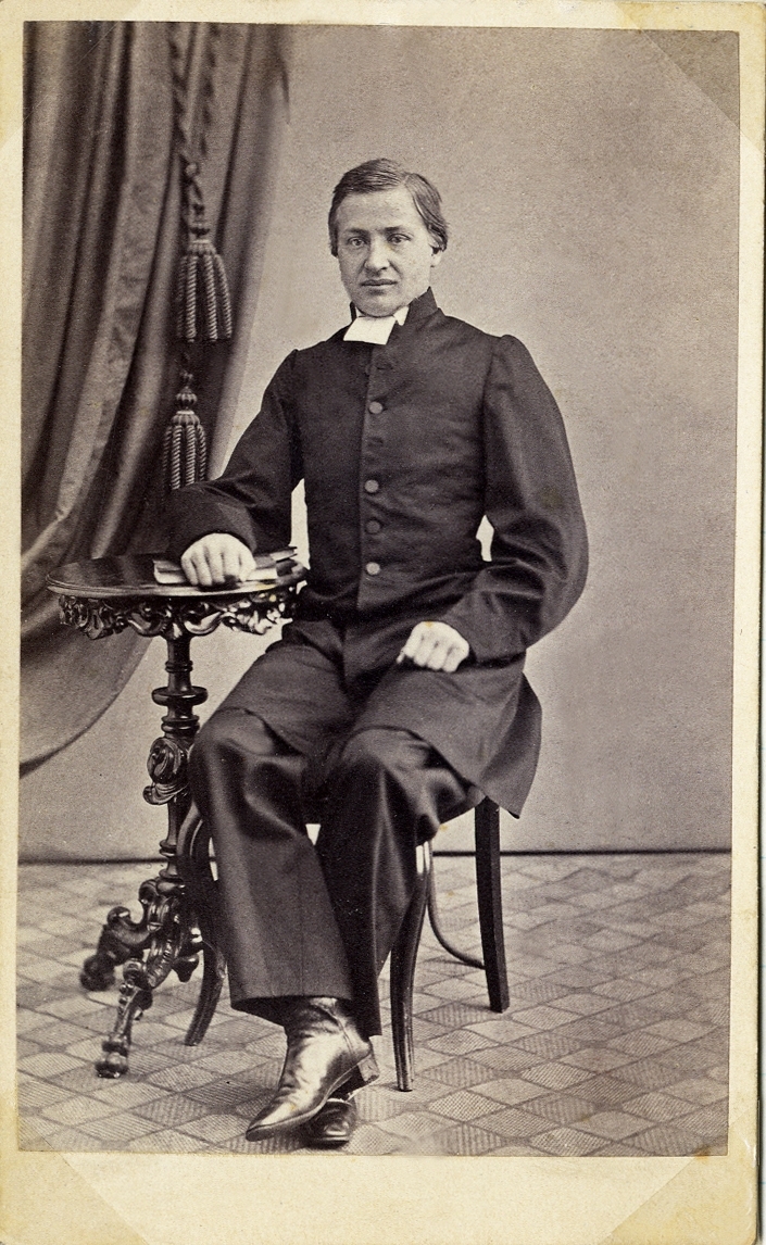 Porträttfoto av en man i prästdräkt. Han sitter på en stol vid ett utsirat pelarbord. I bakgrunden syns ett draperi. 
Helfigur, halvprofil. Ateljéfoto.