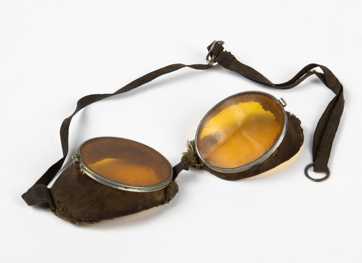 Flygglasögon med gult glas, sidskydd i tyg samt en rem att fästa glasögonen med på huvudet. Runt glasögonens innerkant finns en pälsliknande tyg.
Glasögonen har gått av i mitten och är i två delar.