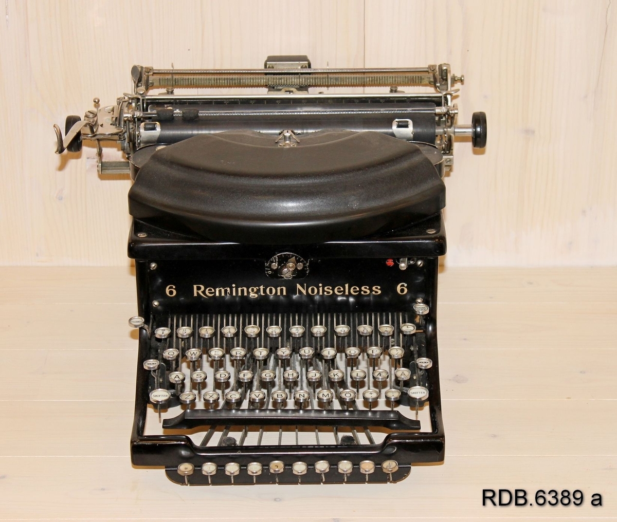 Svart skrivemaskin av merke Remington. Tastene er kvite med svarte bokstaver. På siden av skrivemaskinen har giveren limt på et bilde av og opplysninger om eier og bruker av maskinen.