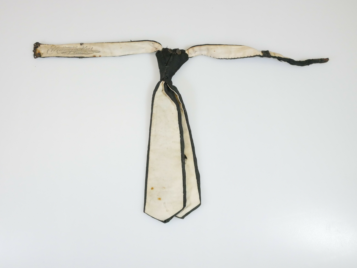 Kort svart slips. Slipsknyten er sydd sammen. De to slipslengdene er like lange og spisse nederst. På baksiden er det et hvitt for. En metallfeste på venstre side. Resten av en metallbit bak på slipsknuten. 