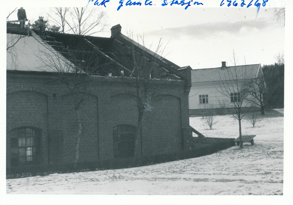Takreparasjon på Nybruket som først huset tresliperi og deretter kraftstasjonen Kistefoss I.  To menn står på taket. Hvite bolighuset Formannsboligen i bakgrunnen.