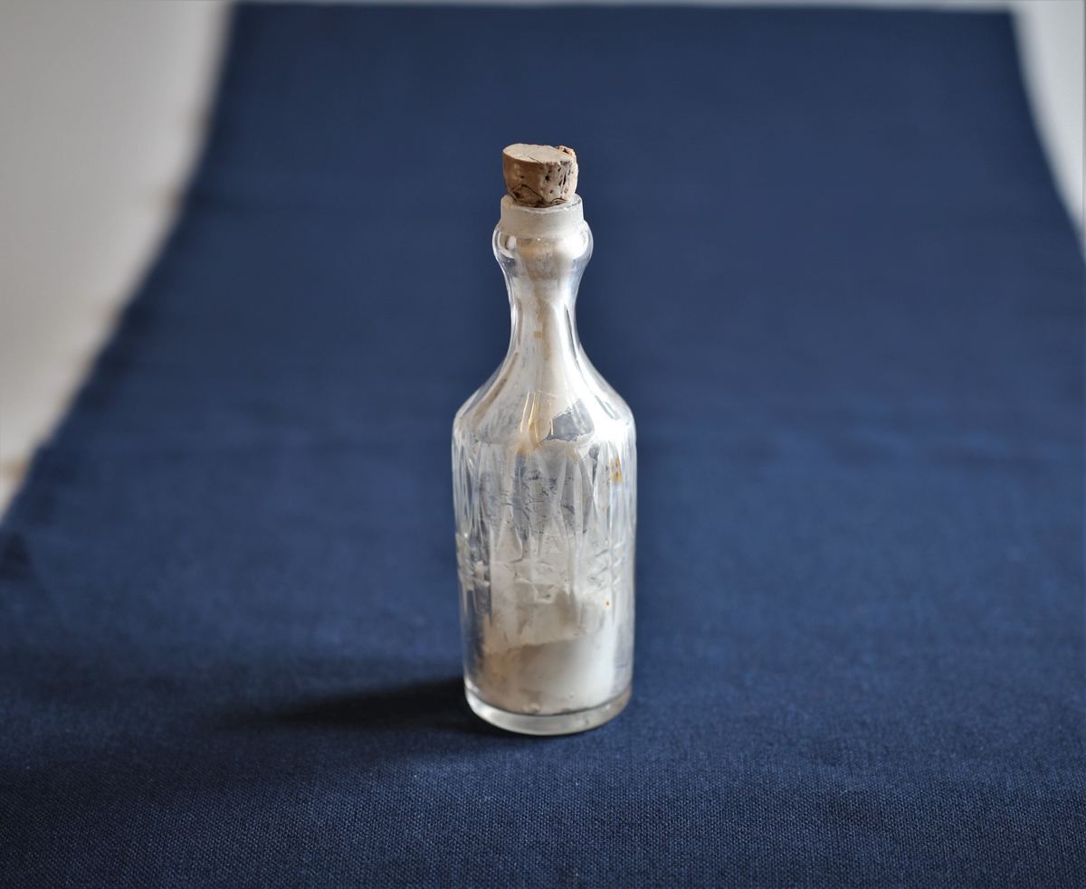 Glassflaske med slepet mønster langs sidene og kork. Den har et grøtaktig, gråkvitt innhold.
