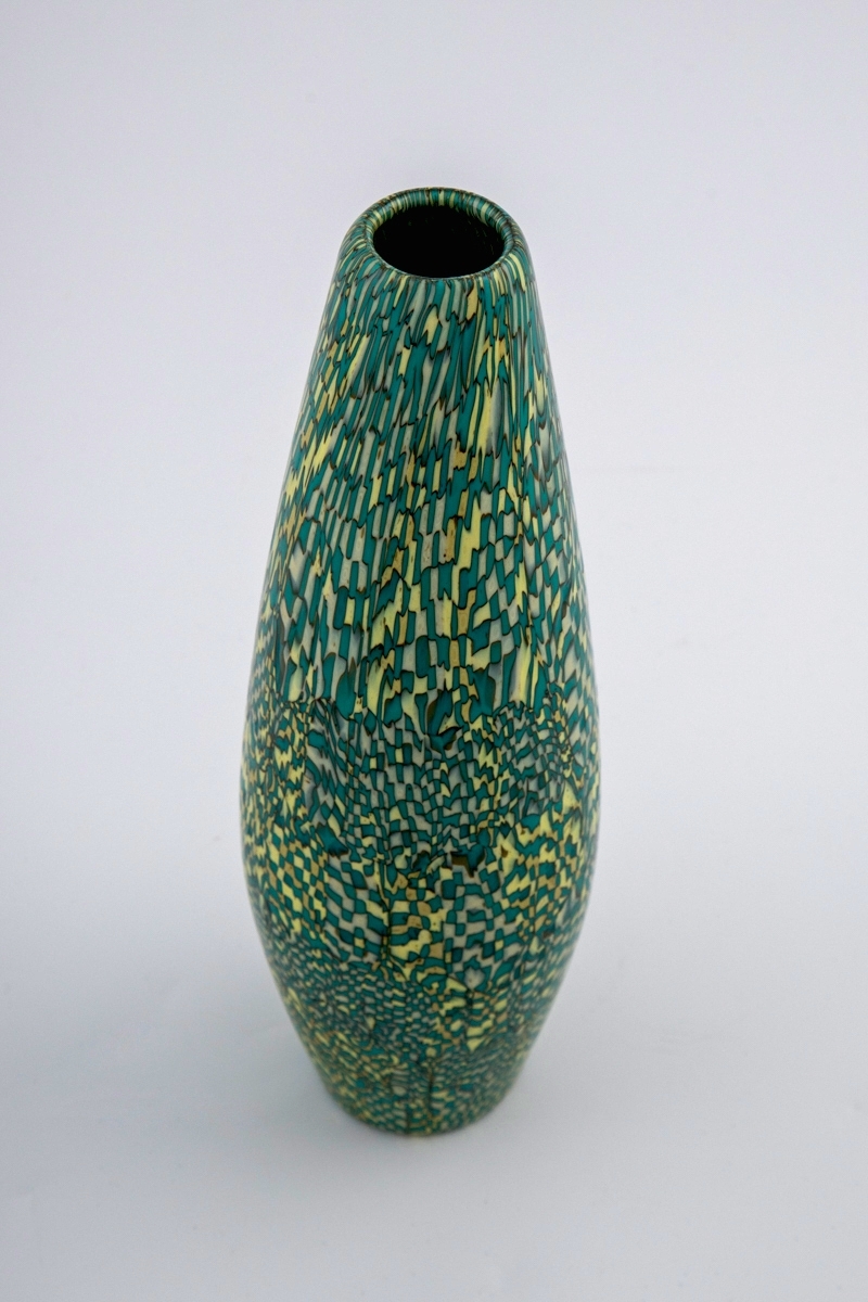 Balusterformet vase i opakt mosaikkglass, bestående av små glasstaver som skaper et geometrisk, uregelmessig mønster i hvitt, gult og turkis, omgitt av sorte konturlinjer. Sirkulær munning og bunn.