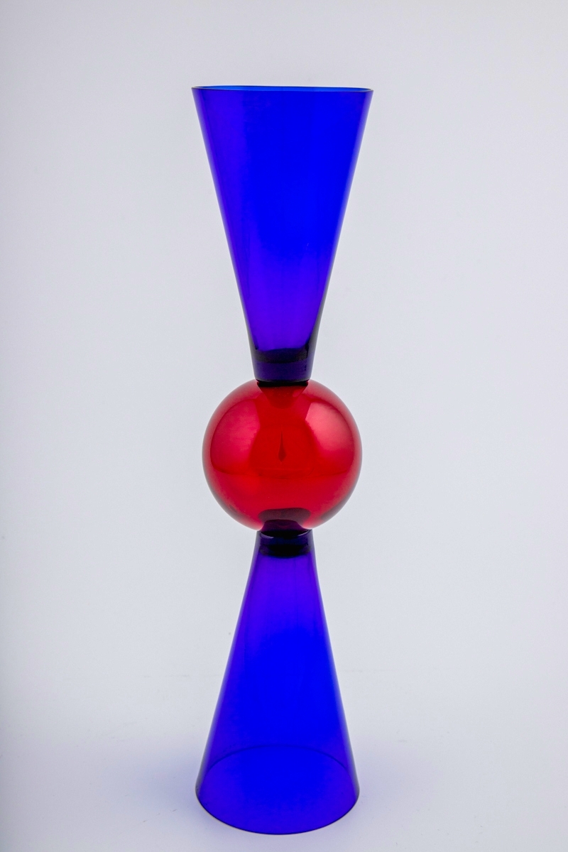 Vinglass utført i farget gjennomskinnelig glass. Blåfarget kupa og fot som er konisk utformet, og smalner mot glassets midtparti som består av en rødfarget kule. Dermed kan begge sidene fungere som fot og klokke.