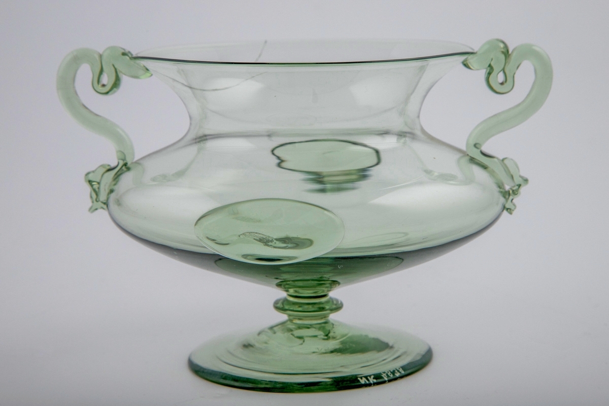 Kraterfromet vase i gjennomskinnelig grønnfarget glass. Kuleformet korpus med to ovale glassknotter, som hviler på en hevet sirkulær fot. Vasen har en lav konisk hals, og to slangeformede glasstaver fungerer som hanker.