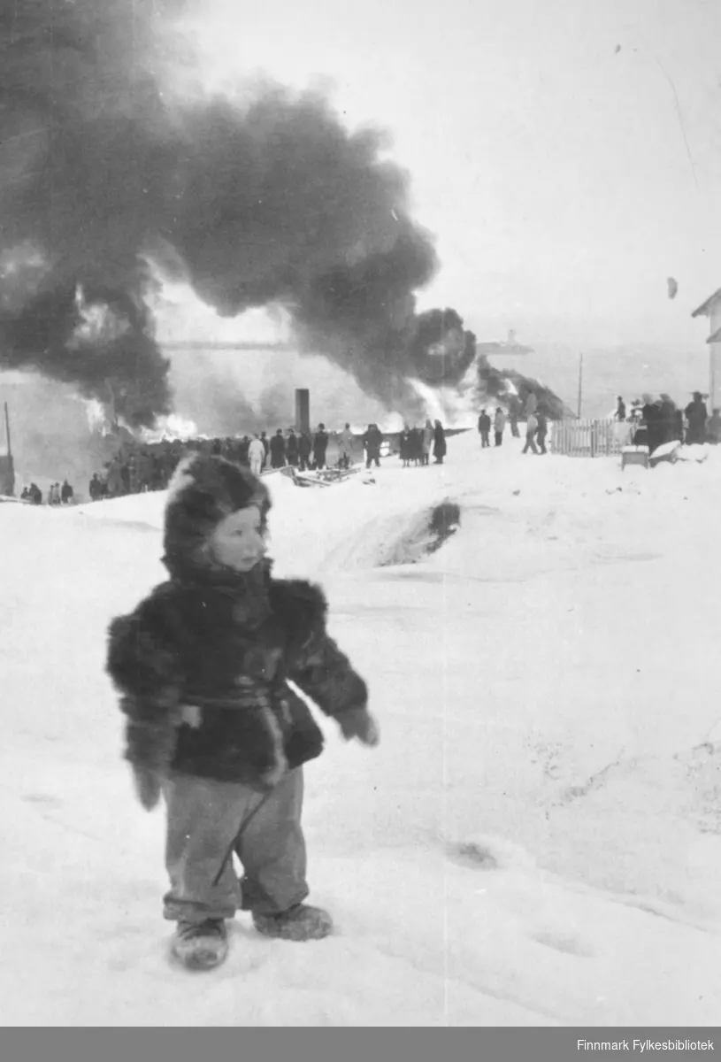 En liten gutt, Jan Erik Sælø,  i bakgrunnen "fiskefabrikk" i Indrebyen i Vadsø brenner, ca. 1955.