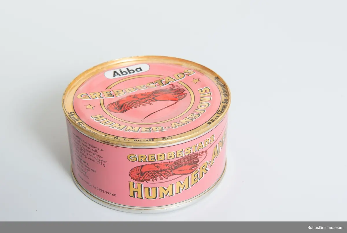 Cylindrisk burk med tryckt dekor på plåten i rosa, rött och guldfärg.
Bild av hummer samt text på lock och sida: "Abba. Grebbestads Hummer-Ansjovis.", 
samt på sidan även innehållsdeklaration och prismärkningskod.

Om givaren: Se UM026667