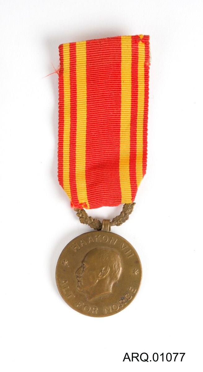 Medalje med Kong Haakon VII på. Tekstilet er rødt med gule loddrette striper. Festenål på baksiden av tekstilet.