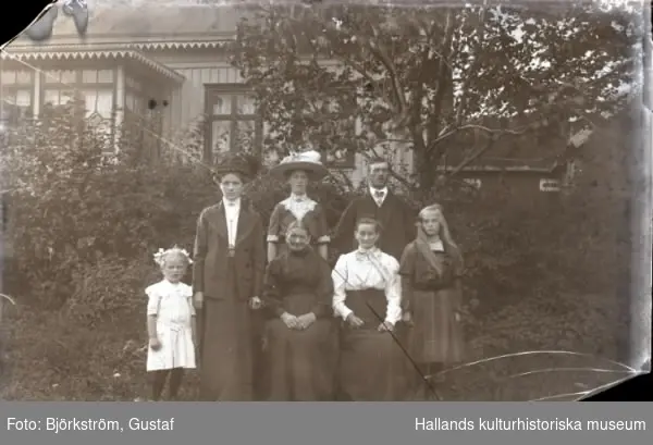 Släktbild med sju personer tagen i Ranelid i trädgården framför bostadshuset med ladugården i bakgrunden. Två kvinnor sitter ned och de två stående kvinnorna bär stora hattar.
(Glasplåten är skadad)