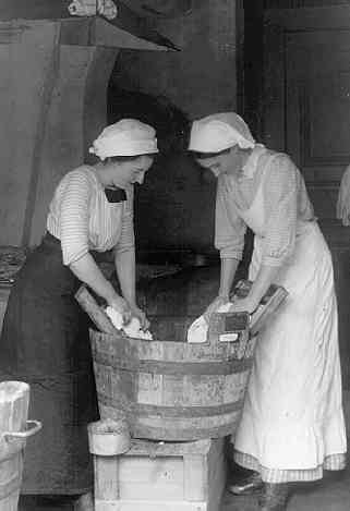Två unga kvinnor med huckle och den ena med ljust förkläde i ett brygghus där de tvättar med tvättbräde i en träbalja. En av kvinnorna är Ester Roman.