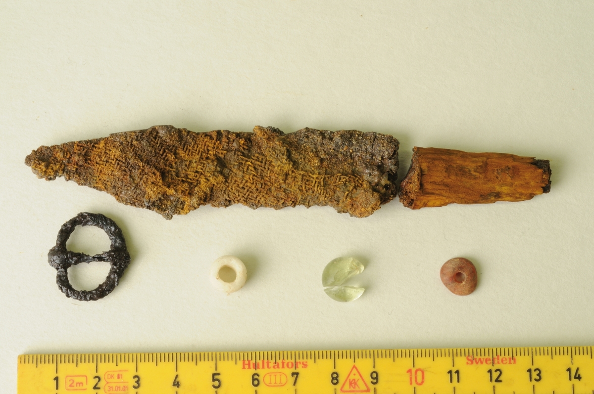 En kniv med rester efter trähandtag, en sölja, en glaspärla, en möjlig spelbricka i glas samt en bärnstenspärla påträffad i en skelettgrav, A 31209, på gravfältet RAÄ Barnarp 29:1, L1974:2724 på Torsvik i Jönköping. I graven påträffades rester efter en individ, troligen pojke, på 12-15 år. Notera resterna efter textil på knivbladet.  Se även bild JMF.00328:13 och JMF.00328-15. Datering 640-685 e.Kr.