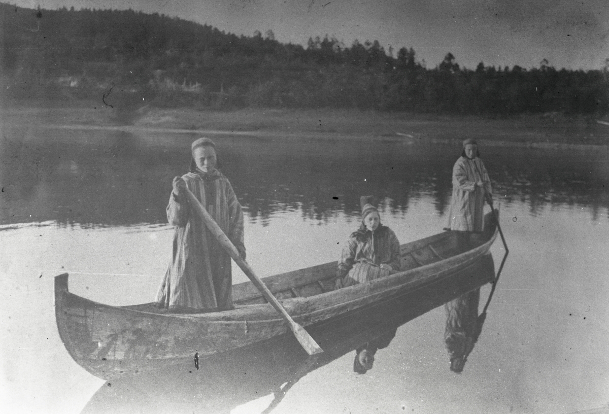 Motiv av tre kvinner i en kano/ elvebåt. Kvinnene har tradisjonelle drakter(kofter) på seg.