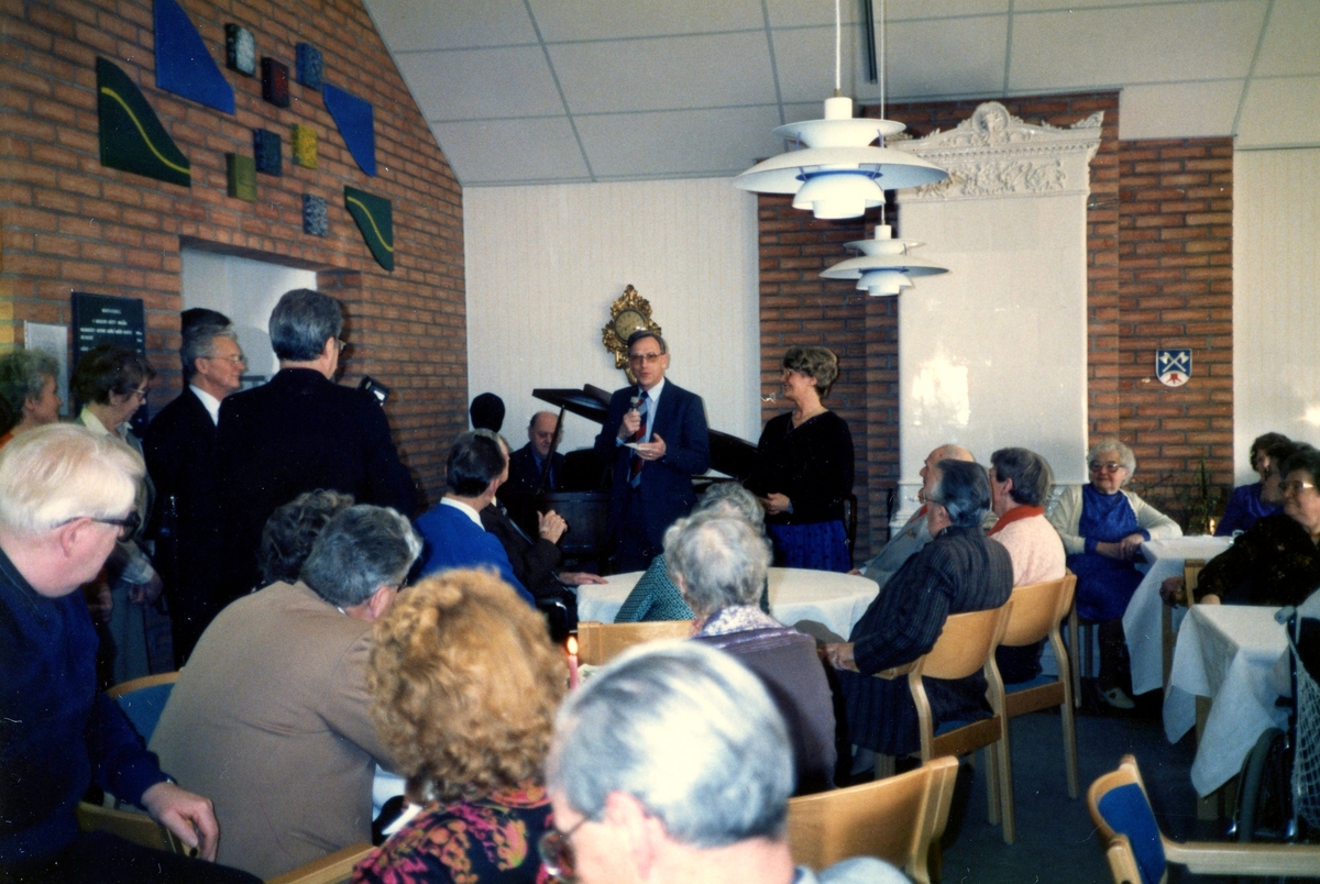 Musikunderhållning i Brattåsgårdens matsal (Streteredsvägen 5) år 1989. Vid flygeln sitter Rutger Milsta (från Lindome), vid mikrofonen står socialchef Bengt Alvekrans och till höger om honom står föreståndare Aina Ekstedt. I bakgrunden skymtar en kakelugn.