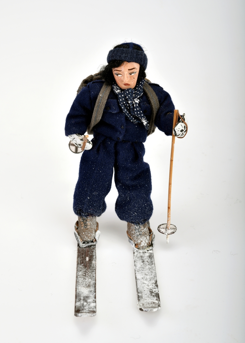 Liten damefigur med ryggsekk, ski på beina og skistaver i hendene.
