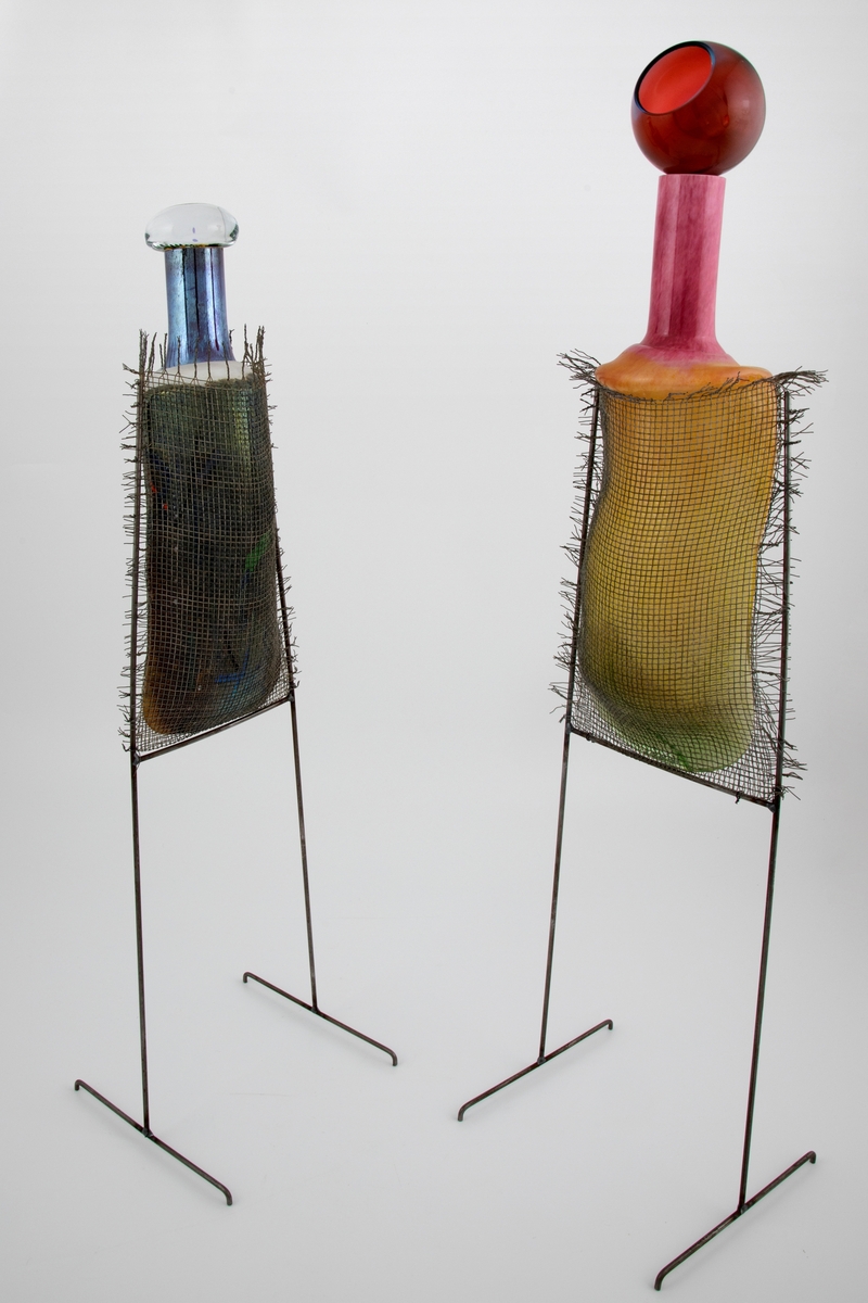 To skulpturer i glass i ulik høyde. Flaskeformet korpus i opakt glass i ulike farger. Sylindrisk hals med tilhørende glasspropp. Begge flaskene er innkapslet i et nettingshylster og bæres av et tobent stativ i metall.