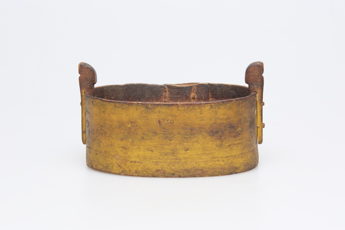 Oval tine (b) med lokk(a), gulmalt, laget av tjukk og bred høvelspon. To oppstandere holder lokket fast. Sviornamenter, tverrsittende håndtak på lokket, der det er påmalt K F D 1872.