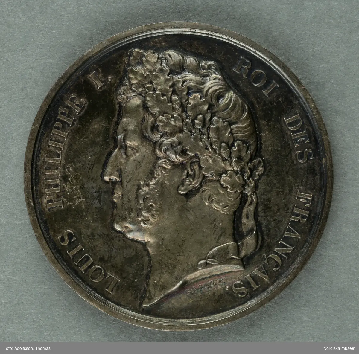 Huvudliggaren.
"Medalj. I hvit metall. Åtsidan: 'Louis Philippe I Roi des francais'. Bröstbild v.o. Under bilden: 'BARRE F.T [t:et snett ovanför F.et]. Frånsidan öfverst: 'Ville d'Arras'. Inom en eklöfskrans: 'Exposition de 1838. A. M [efter m;et svårtytt tecken] Wickemberk peintre à Paris'. Nederst utom kransen: 'Dep .T [t:et ovanför punkten] du Pas-de-Calais.'. 
Diam lin.
G. fr basaren f. Skdin. etn. saml. [=för Skandinaviska etnografiska samlingens] byggnadsfond, gm. fru Olivecrona f. Roos, i apr. 1877."