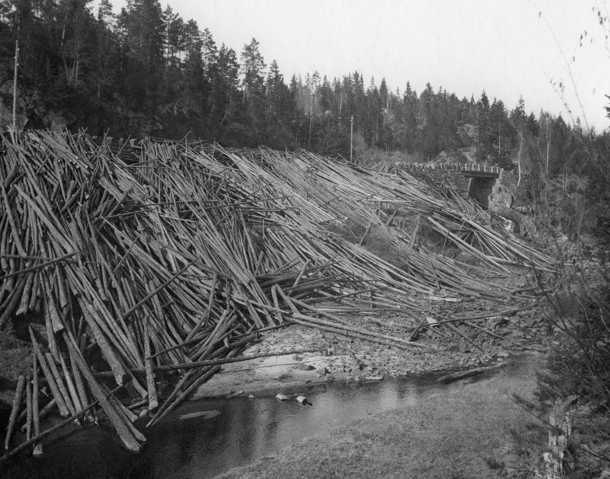 Tømmer i skråning mellom bilveg og vassdrag i Bamble i Nedre Telemark.  Det barkete tømmeret er åpenbart veltet over stabbesteinene langs vegens ytterkant og utfor den forholdsvis bratte skråningen, der fløterne antakelig måtte hente det for å kunne få slått det uti elva ved høvelig vannføring.  Bildet skal være tatt våren 1921.  1920-21 var et driftsår i skogbruket og skogindustrien som ble preget av stor etterspørsel og intens drift som ble avløst av et krakk.  Etterspørselen og prisene i tømmermarkedet hadde vokst gjennom flere år, og aktiviteten i næringa var stor.  Industrien inngikk kontrakter om store tømmerkjøp, og skogeierne organiserte storstilte hogster.  Tømmeret ble transportert fram til fløtingsvassdragene, men da våren kom hadde prisene falt.  Mye av inntektsgrunnlaget for den norske skogindustrien og bedriftenes grossister i utlandet falt bort, og mange hadde vanskeligheter med å betale skogeierne de avtalte prisene, og måtte be om henstand.  Fullt oppgjør ville blant annet ha veltet storbedriften Union Co. i Skien.  Høykonjunkturen ble avløst av kraftig prisfall og langvarig depresjon.  Dette førte til konkurser.  Mange av skogeierne gikk konkurs, og det gjorde også Skogeierbanken og andre kredittinstitusjoner som hadde skogeiere og skogindustri som viktige kundegrupper.  Med den labre etterspørselen etter tømmer utover i 1920- og 30-åra var det neppe var slike mengder tømmermengder i vegskråningene langs vassdragene i Bamble. 

At det lå slike tømmermengder som fotografiet viser langs elvene våren 1921 skyldtes ikke bare stor hogstaktivitet - det skyldtes også at det knapt ble noen vårflom, slik at det var vanskelig å få fløtet tømmeret med vanlig effektivitet.
Dette fotografiet ble publisert i årsmeldinga til fylkesskogmester Gisle Midttun for driftsåret 1921.  Fylkesskogmesterens beskrivelse av bakgrunnen for at det lå slike tømmermengder langs elvebreddene er gjengitt under fanen «Opplysninger».