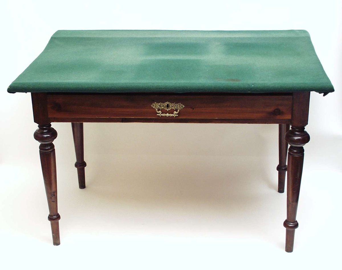 Skrivebord i beiset gran med dreide profilerte ben. Bordet har en skuff med trekkbeslag i sargen. Overplaten har bakstykke og er trukket med grønt tøy. Den opprinnelige bordplaten er 64,5 bred.