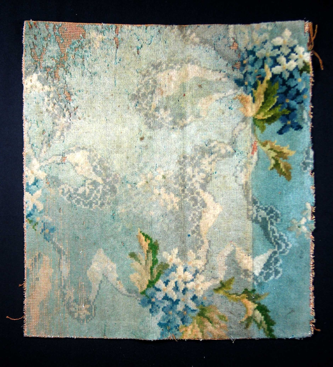 Fragment av golvteppe i ullfloss. Motivet er blomster og bladverk i nyanser av blått, grønt, beige og hvitt. Maskintilvirket.
