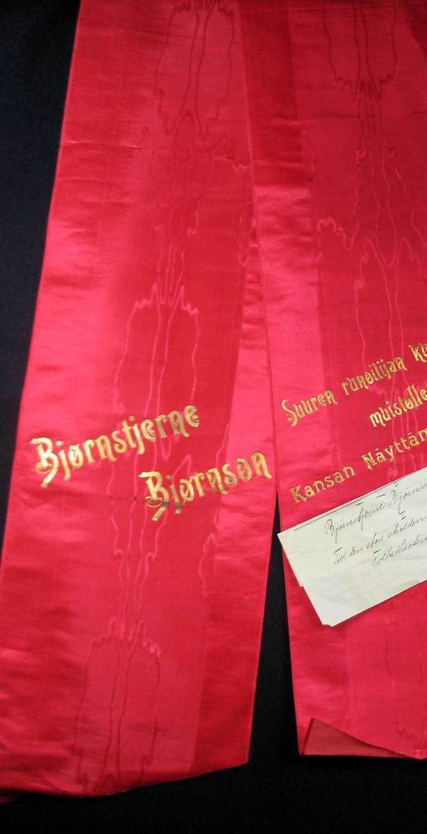 Begravelsessløyfe i rød mønstret silke med tekst i gull.