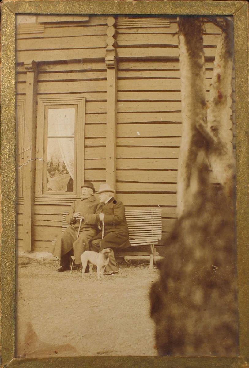 To menn på en benk foran en tømmerbygning. En hund ved siden av.