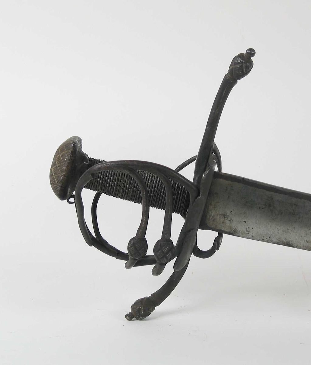 Pyramideformet knapp med strekdekor. Asymmetrisk kurvfeste med to sidebøyler( øverste avbrukket )med riflede fortykkelser på midten. mellom håndbøylen og kurvens ytterkant. Innsiden har en  sidebøyle. Kurvens basis er på utsiden en kvadratisk parérplate med to hjerteformede gjennombrytninger. På innsiden er det en klingebrekker og tommelbøyle. Opp og nedbøyde parérstenger med riflede fortykkelser på endene.
Tregrep med nyere jerntrådvikling.
Enegget krum klinge med 3 hull ipå klingspissen og en smal hulslipning på ryggsiden.Tre smedmerker på venstre side. 