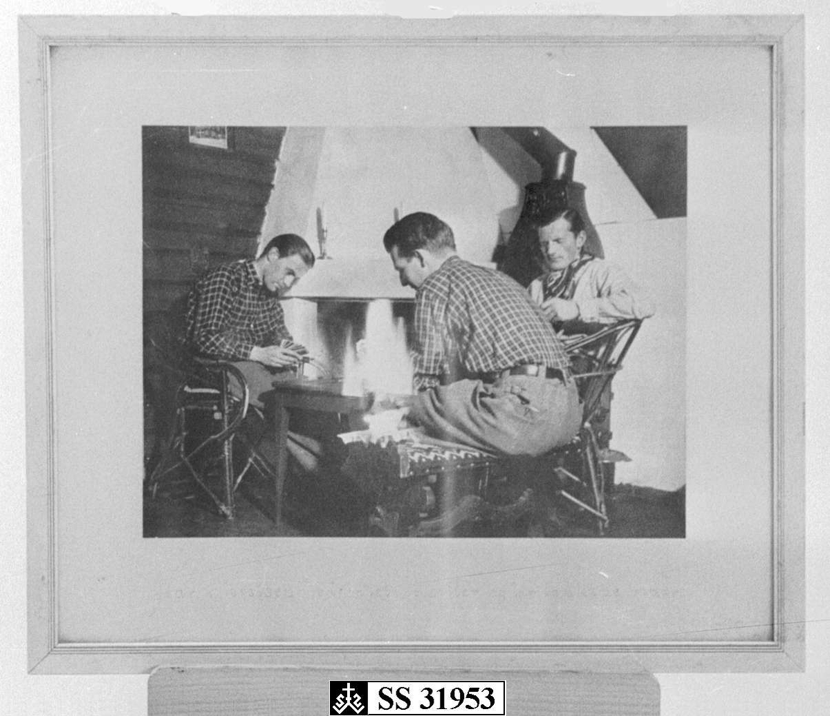 Tre menn ved peisen som spiller kort.