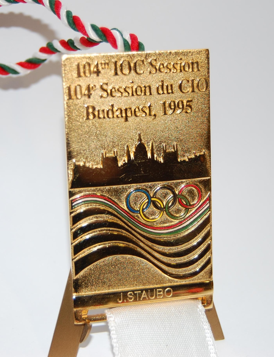 Gullfarget jakkemerke med motiv av de olympiske ringene i farger og Budapest. Til merket er det festet et hvitt bånd.