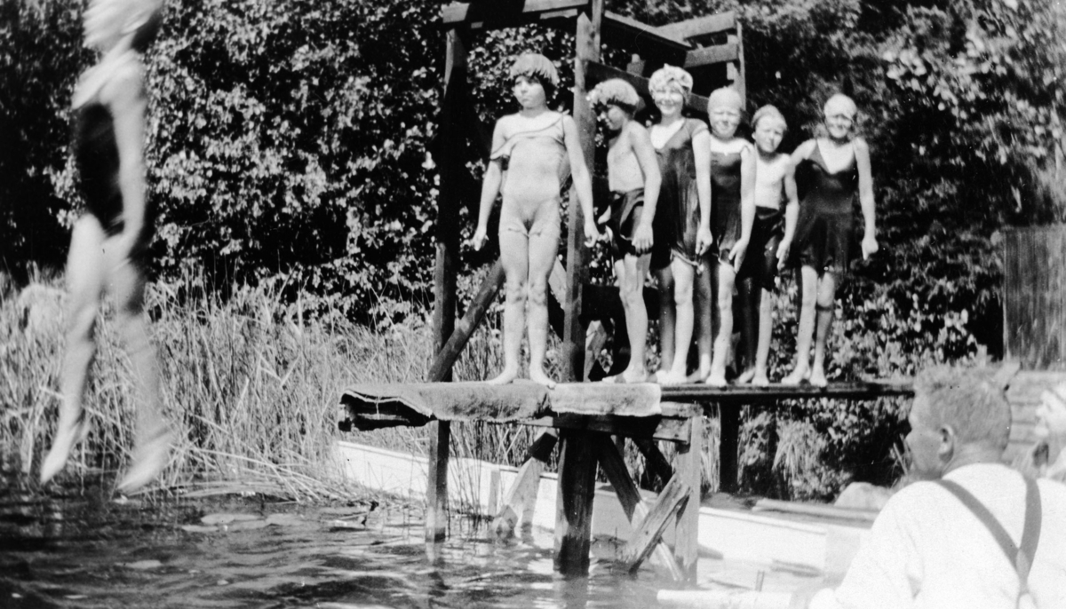 Bading i Langtjernet, østsiden ved Vangen. Barn iført badedrakter og badehetter hopper fra stypebrettet. En voksen mann er tilskuer.