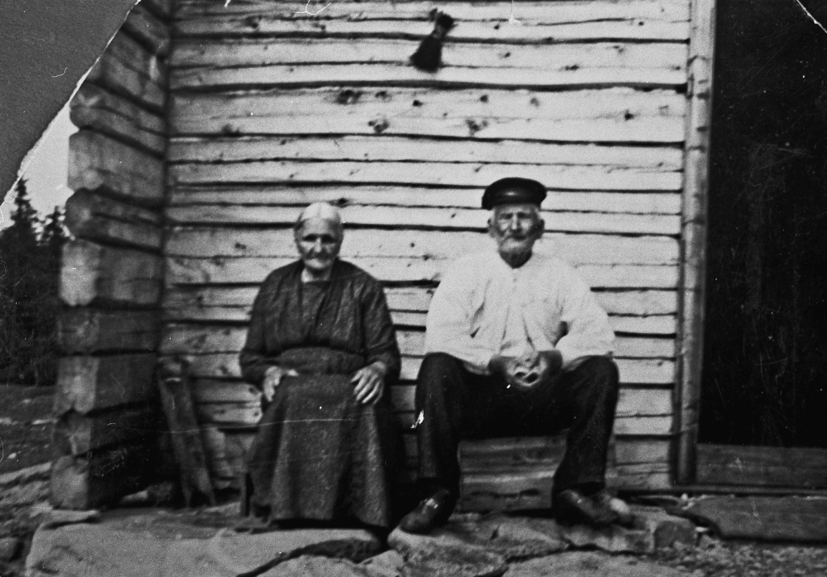 Eldre kvinne og mann utenfor et tømmerhus.
