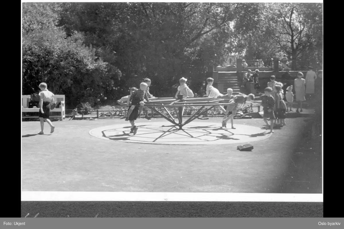 Uranienborgparken med lekeplassen syd for kirken. Ligger mellom skolen og kirken. Opparbeidet i 1922. Barn i lek på karusell. Trappegang opp mot kirken. Sommer.