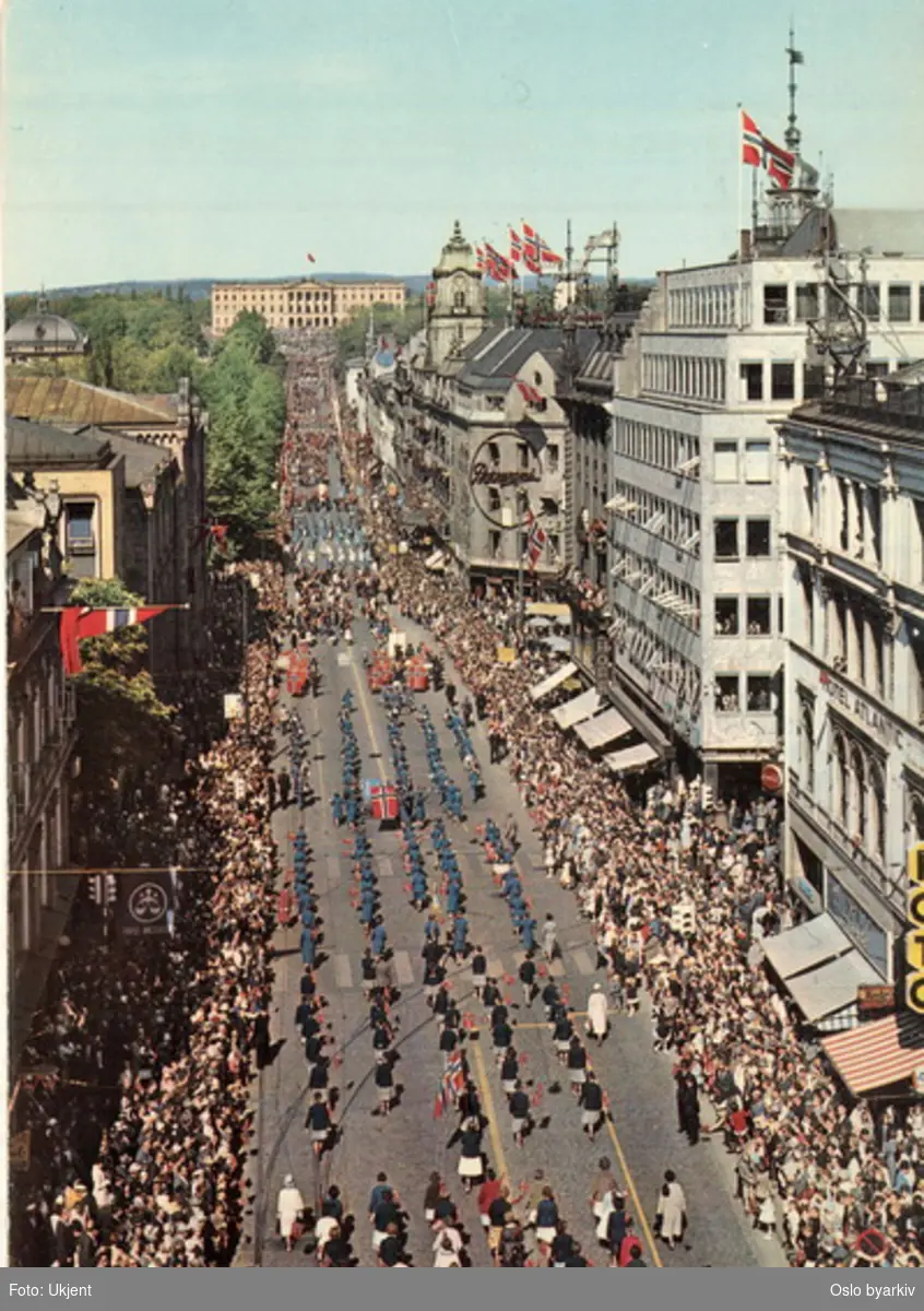 17. mai-tog på Karl Johans gate, sett fra Egertorget til Slottet. Flagg, musikkorps, faner, tilskuere. Kontor- og forretningsgårder. Postkort.