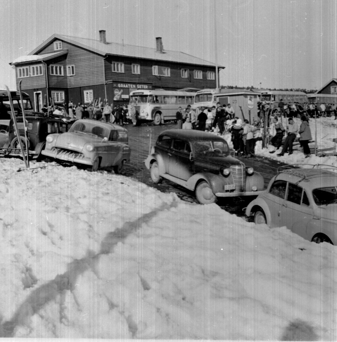 Sjusjøen, bussavgang fra Graaten til Hamar og Lillehammer. Mye folk, påsketrafikk. Bilene i forgrunn er fra venstre ukjent merke ca. 1932, E-54 Opel Rekord varebil 1953, Den mørke bilen, D-3535, er en Chevrolet 1938,  Morris Minor.