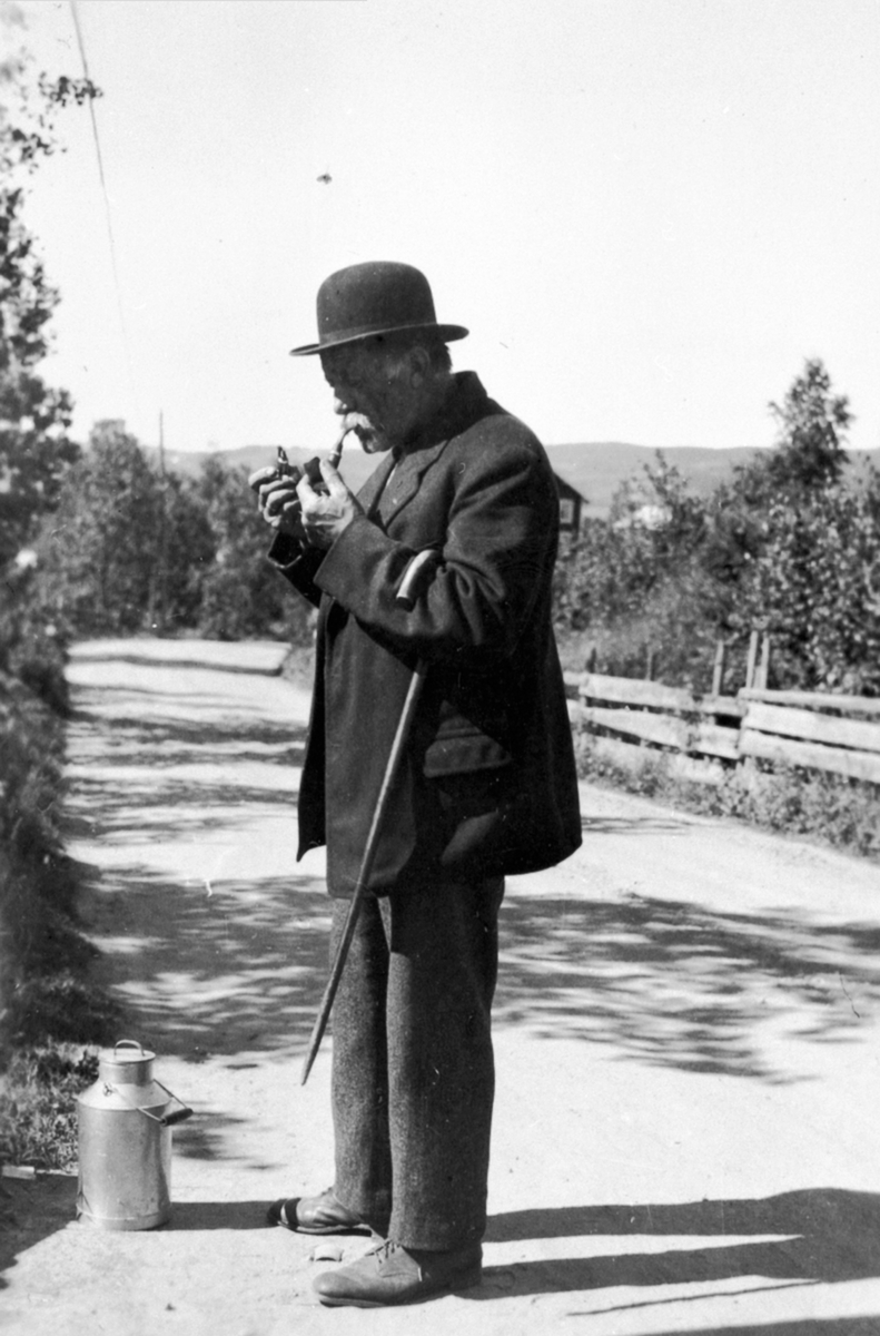 Anton Syverstuen med hatt, stokk og melkespann, tenner pipa.