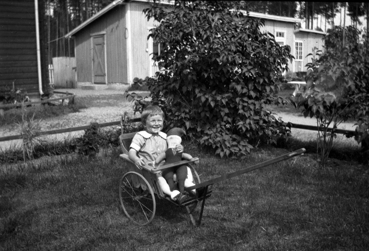 Barn med nissedokke sitter i en slags vogn.
Bildet er tatt på Nortun, gnr. 31, bnr. 178 i Elverum. Barnet er Odd Skibness (1927-1997). Odd vokste opp på Nordtun.