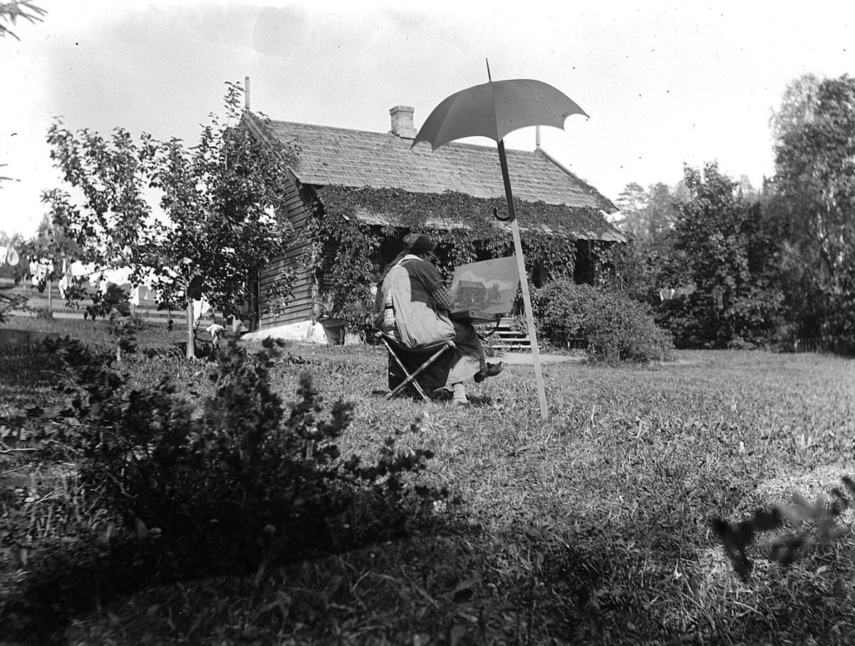 Kvinne under parasoll sitter og maler i hage. Aagot Olssøn, Kvalstua, Helgøya. 