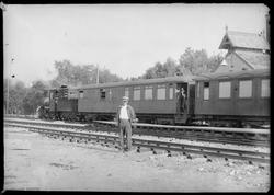 Tog med lokomotiv og vogner står foran stasjonsbygninga på R