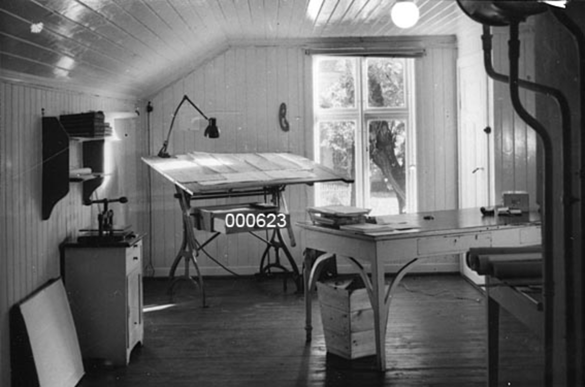 Interiør fra laboriatoriebygningen til A/S Buskerud Papirfabrikk på Åssida i Drammen (den gang i Lier kommune).  Laboratoriet lå i et ombygd bryggerhus på Bergskaug, et gammelt gardstun som lå vis a vis fabrikken.  Fotografiet viser et rom med faspanelte vegger og tak, som er malt i lyse farger.  Taket er skrådd mot husets langvegger.  I rommet står to bord - et flatt arbeidsbord med en del papir på og med papirkorg under, og et tegnebord med skrå plate med egen skrivebordslampe og med underliggende skuff.  Ved langveggen til venstre står en liten kommode med ei presse på toppflata.  Over denne henger ei lita hylle.  Rommet får lys fra et krysspostvindu på endeveggen og fra en kuleformet kuppel i taket. 

Laboratoriebygningens eksteriør er avbildet på SJF-F. 000617.  SJF-F. 000619 - SJF-F. 000622 viser andre deler av interøret i bygningen. 