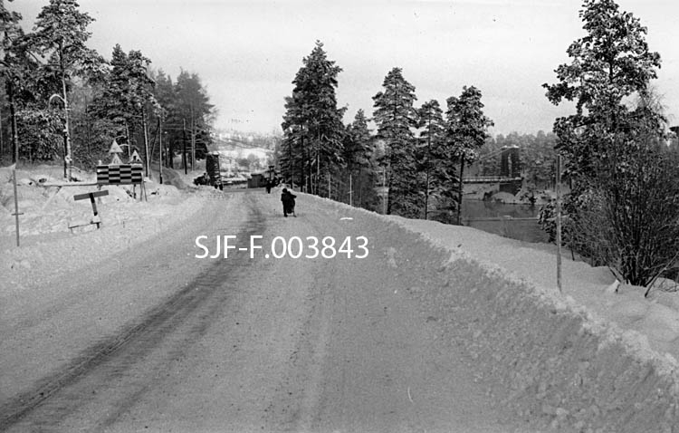 Fra fylkesvegen mellom Geithus og Vassbunn på Modum i Buskerud.  Bildet er tatt mot nord.  I bakgrunnen ser vi Geithus bru, lokalt kalt "Nybrua, til tross for at den ble åpnet så tidlig som i 1909.  Jernbanebrua på Randsfjordbanens sidespor fra Geithus stasjon til Drammenselvens Papirfabrikker er nemlig enda eldre.  Jernbaneforbindelsen ble etablert da fabrikken ble satt i virksomhet i 1874, men jernbanebrua (som vi knapt skimter på dette fotografiet) er seinere ombygd.  Dette fotografiet er tatt vinteren 1962.  Vegen var brøytet for biltrafikk, men da bildet ble tatt var et barn med spark eneste trafikant. 