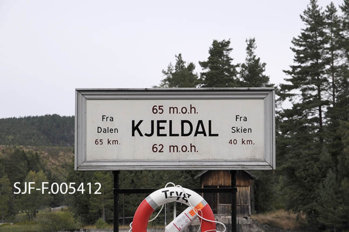 Innrammet skilt på påle ved Kjeldal sluse i Bandak-Norsjø-kanalen.  Skiltet forteller at sluseanlegget ligger 62-65 meter over havet.  Differansen på tre meter refererer til løftehøyden, som i det ene slusekammeret her ved Kjeldal er på tre meter.  Det samme skiltet forteller også at Kjeldal sluse ligger 40 kilometer fra Skien i den ene enden av det kanaliserte vassdraget og 65 kilometer fra Dalen i Tokke, som var det innerste anløpsstedet for båtene som gikk på det kanaliserte vassdraget.  Under skiltet med denne informasjonen henger ei redningsbøye. 