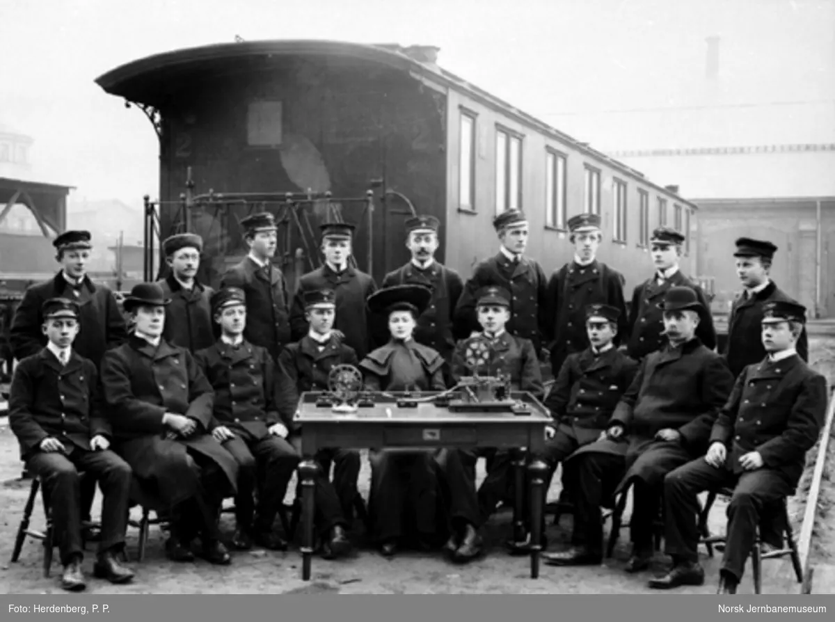 Gruppebilde med 18 telegrafistelever foran personvogn litra Bo nr. 905 og bak et bord med telegrafiapparater