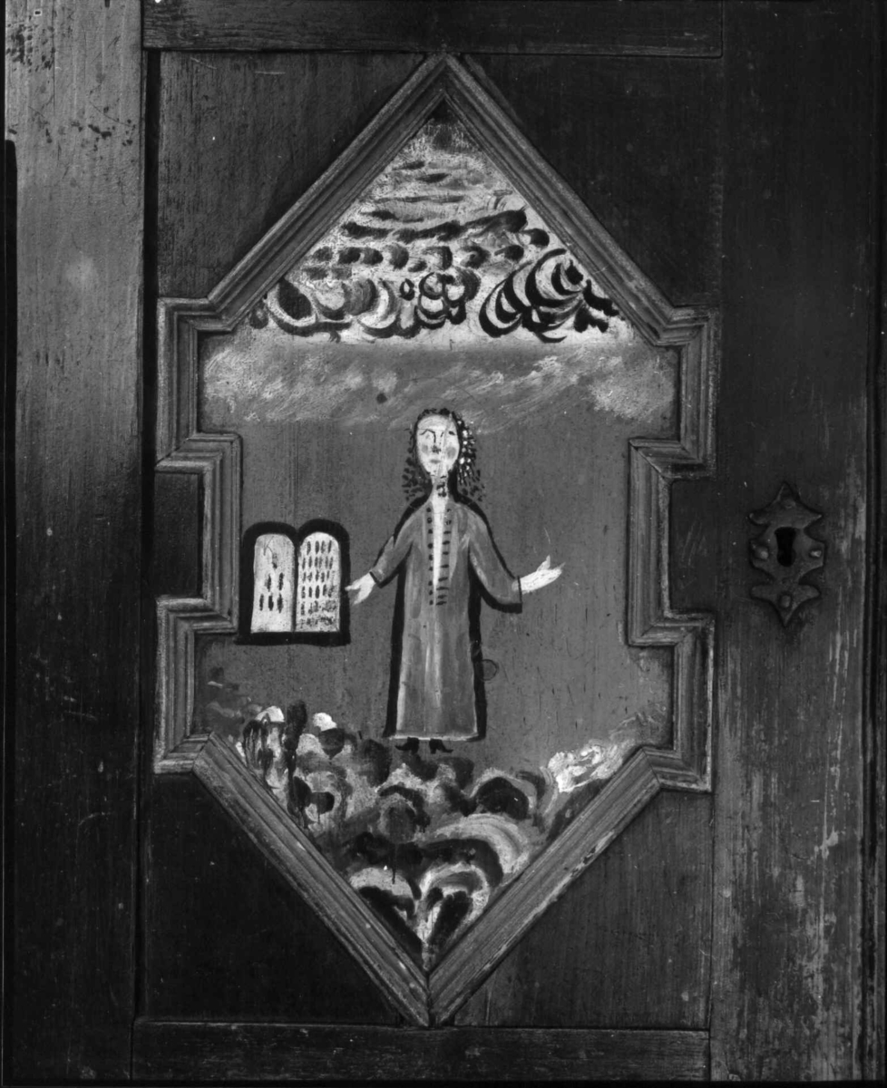 Skap, framskap i en del, to dører oppe og nede. Bibelske motiver; Moses og de ti bud. Gud på en sky, en brennede sky eller busk, en mann som har mistet en sko og en stokk.