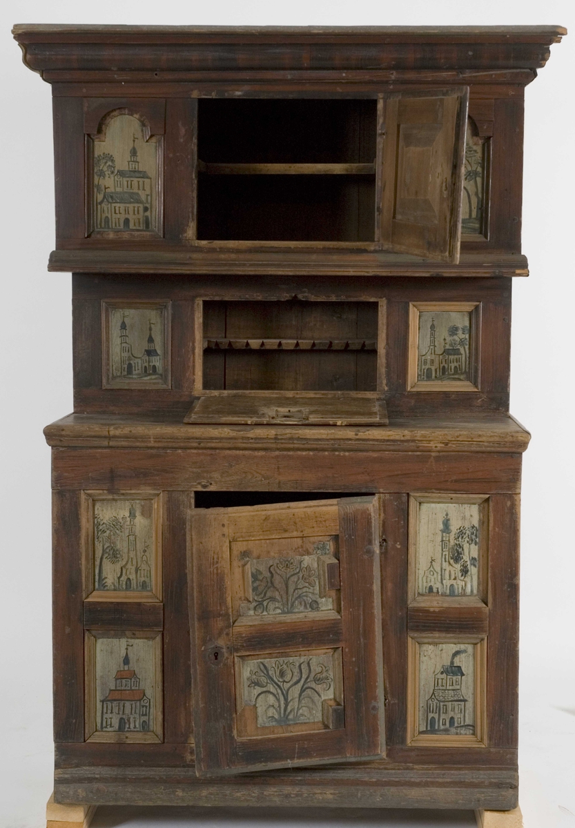 Framskap i en del med en dør nede, en  oppe og en liggende i midten. Dørene har malte blomstermotiver mens de 8 sidefeltene har malte bygninger.