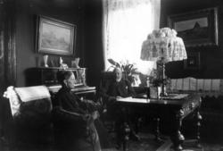 Interiør, ukjent sted. 1908-1910. Stue. Kvinne og mann ved b