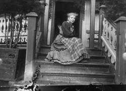 Drakt 1908-10. Kvinne sittende i en trapp. Veranda i balgrun