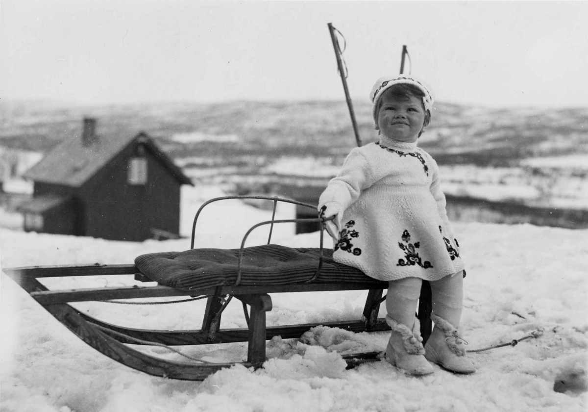 En liten jente med hvit, brodert strikkekjole, votter og lue i samme mønster sitter på kjelke. I bakgrunnen sees et bolighus og en snødekt dal.
